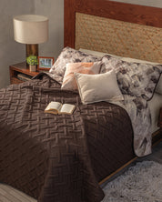 A cozy bedroom features the Juego de Edredón Ligero Coffee by Intima Hogar, patterned pillows, and an open book on the bed. A wooden nightstand with a lamp, potted plant, and photo sits beside the bed’s wooden headboard.