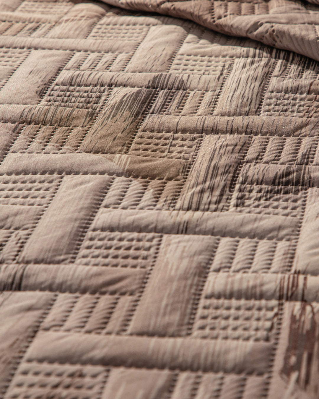 A close-up of the Juego de Edredón Ligero Coffee by Intima Hogar displays its quilted geometric square pattern in brown, highlighting a large visible wet stain on the fabric.