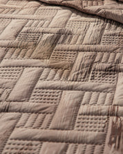 A close-up of the Juego de Edredón Ligero Coffee by Intima Hogar displays its quilted geometric square pattern in brown, highlighting a large visible wet stain on the fabric.