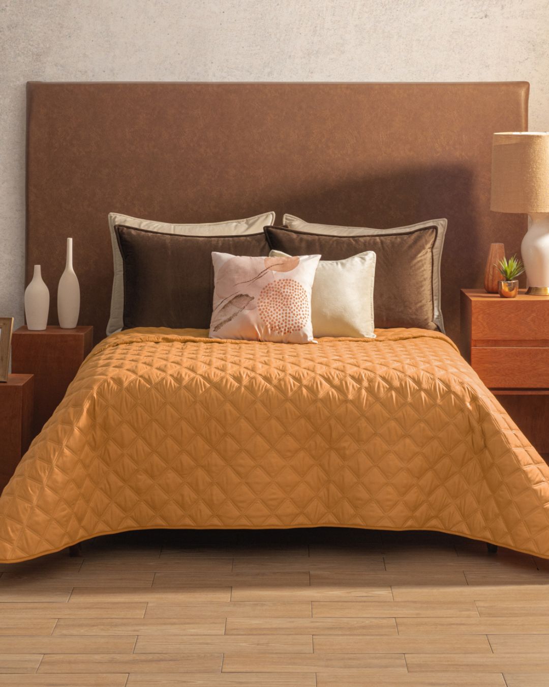A neatly made bed features the Intima Hogar Juego de Edredón Ligero Destello in tan, three decorative pillows, and a cushioned brown headboard. Matching wooden nightstands with lamps and vases add a subtle shine to this cozy contemporary room with wood floors.
