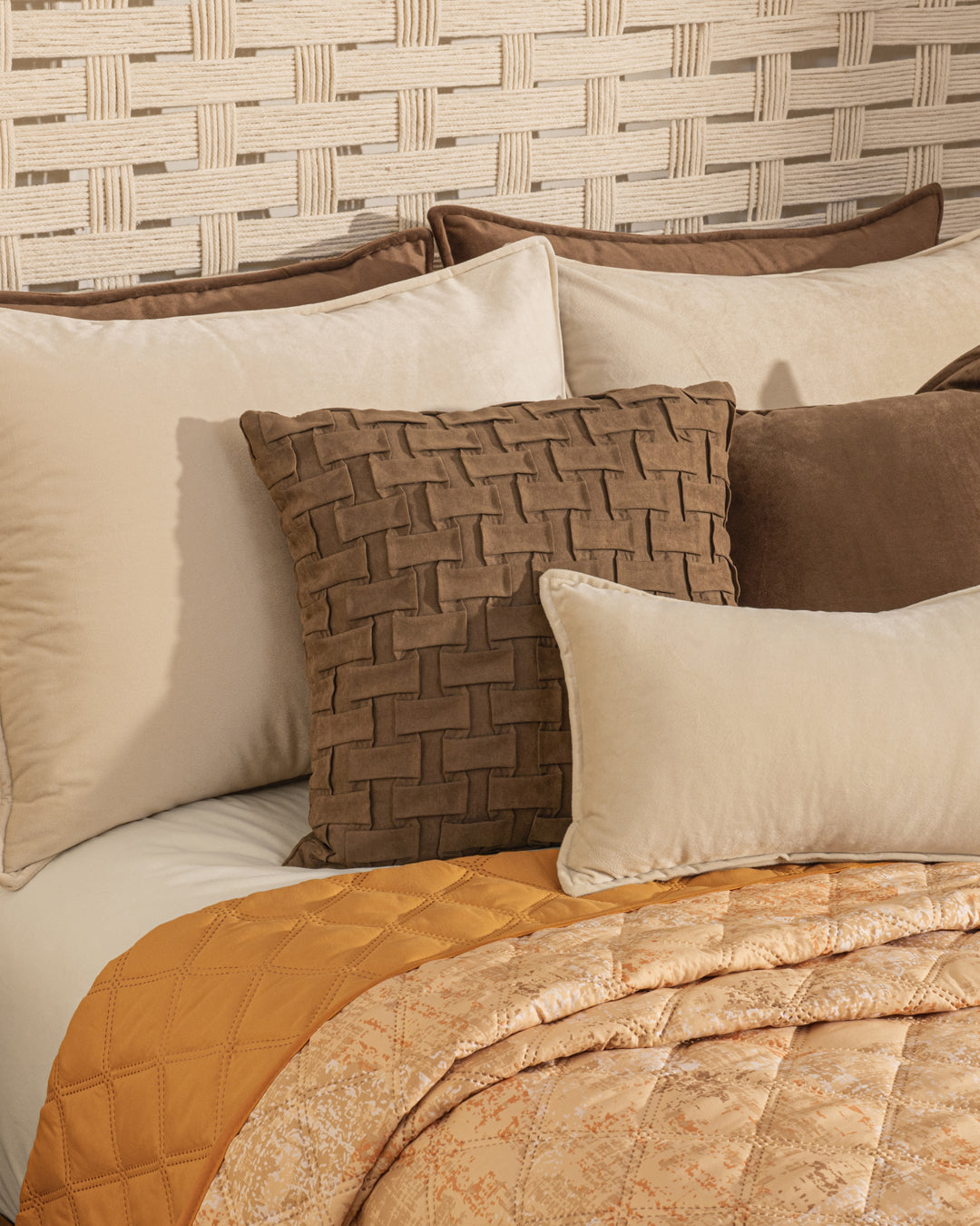 A neatly made bed features the Intima Hogar Juego de Edredón Ligero Destello, beige and brown pillows, a textured throw pillow, and a gold quilted blanket, all set against a woven-patterned beige headboard.