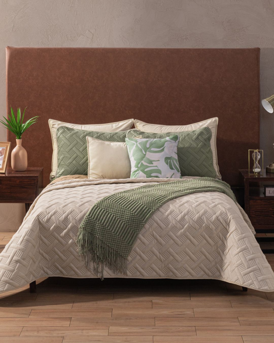 A neatly made bed features the Intima Hogar Juego de Edredón Ligero Esperanza, a brown headboard, green and cream pillows, and a green throw. Wooden nightstands with lamps, books, and plants flank the bed on light wood flooring.