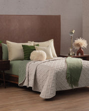 A neatly made bed with the Juego de Edredón Ligero Esperanza by Intima Hogar, green and cream fundas decorativas, and a green throw sits beside a wooden nightstand with a lamp, vase of dried flowers, and book in a cozy, diseño elegante bedroom.