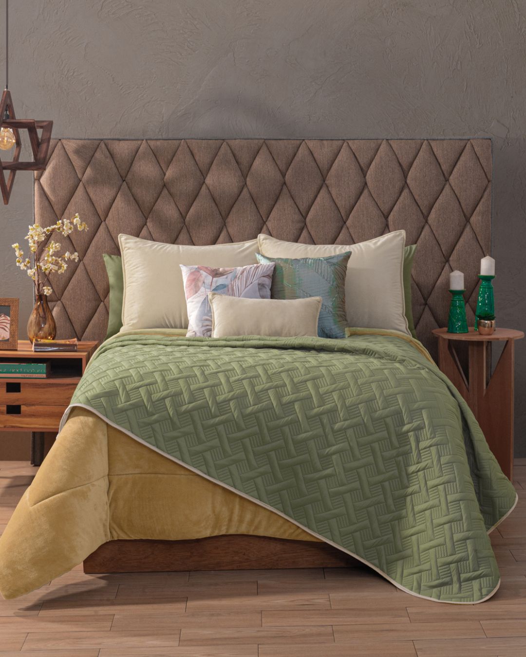 A neatly made bed features the Intima Hogar Juego de Edredón Ligero Esperanza with a tufted headboard, gold blanket, and decorative pillows. Two wooden nightstands with lamps, vases, and decor sit on each side atop a wooden floor.