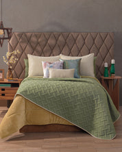 A neatly made bed features the Intima Hogar Juego de Edredón Ligero Esperanza with a tufted headboard, gold blanket, and decorative pillows. Two wooden nightstands with lamps, vases, and decor sit on each side atop a wooden floor.