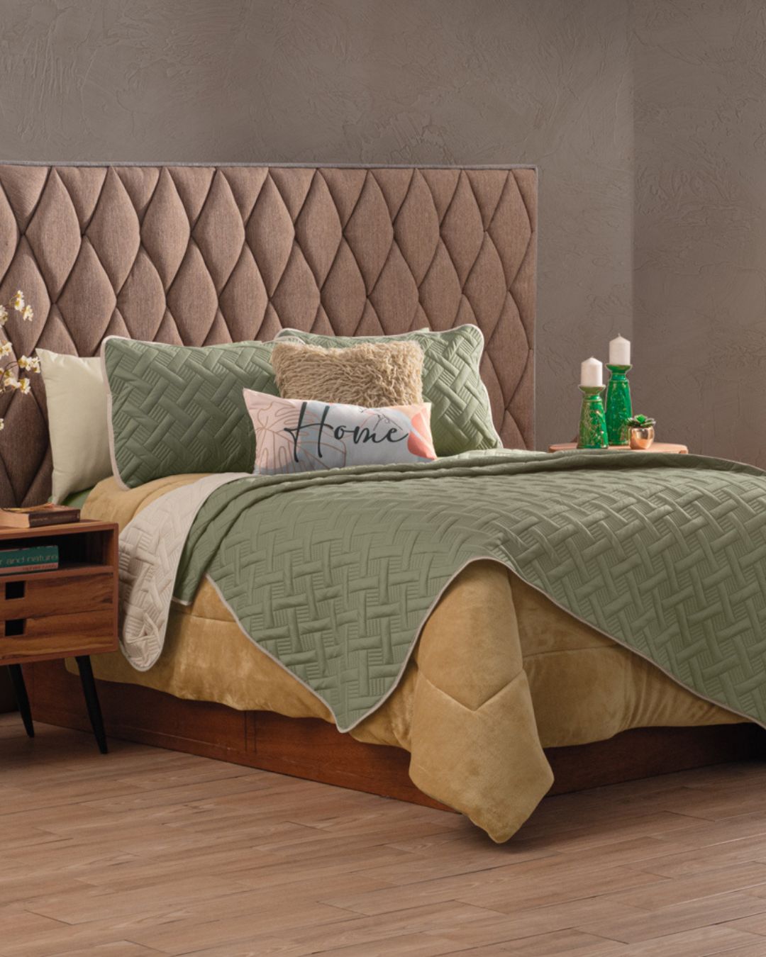 A neatly made bed features the Intima Hogar Juego de Edredón Ligero Esperanza, sage green textured coverlet, assorted decorative pillow covers (one reads "Home"), and a tall padded headboard. Wooden nightstands with books, candles, and green decor flank the bed.