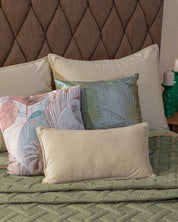 A neatly made bed features the Juego de Edredón Ligero Esperanza by Intima Hogar, with a green quilted blanket, four decorative pillows in soft hues, a brown tufted headboard, plus candles and green glass decor on the side table.