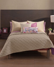 A neatly made bed features the Intima Hogar Juego de Edredón Ligero Espíritu, beige and purple pillows with fundas decorativas, a "Tropical" decorative pillow, a dark wood headboard, and nightstands with a lamp, plant, and framed photo.
