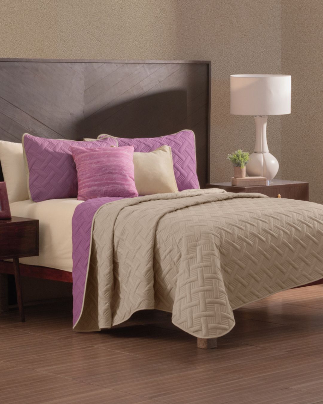 A neatly made bed showcases the Juego de Edredón Ligero Espíritu by Intima Hogar in beige and purple, with matching pillows and a pink accent pillow. A wooden nightstand with a white lamp and plant sits beside it on a wood floor.
