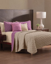 A neatly made bed showcases the Juego de Edredón Ligero Espíritu by Intima Hogar in beige and purple, with matching pillows and a pink accent pillow. A wooden nightstand with a white lamp and plant sits beside it on a wood floor.