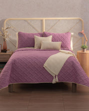 A neatly made bed showcases the Juego de Edredón Ligero Espíritu by Intima Hogar in lavender, with matching and beige accent pillows. A cream tassel throw is draped on the bed, and nightstands with decor flank each side.