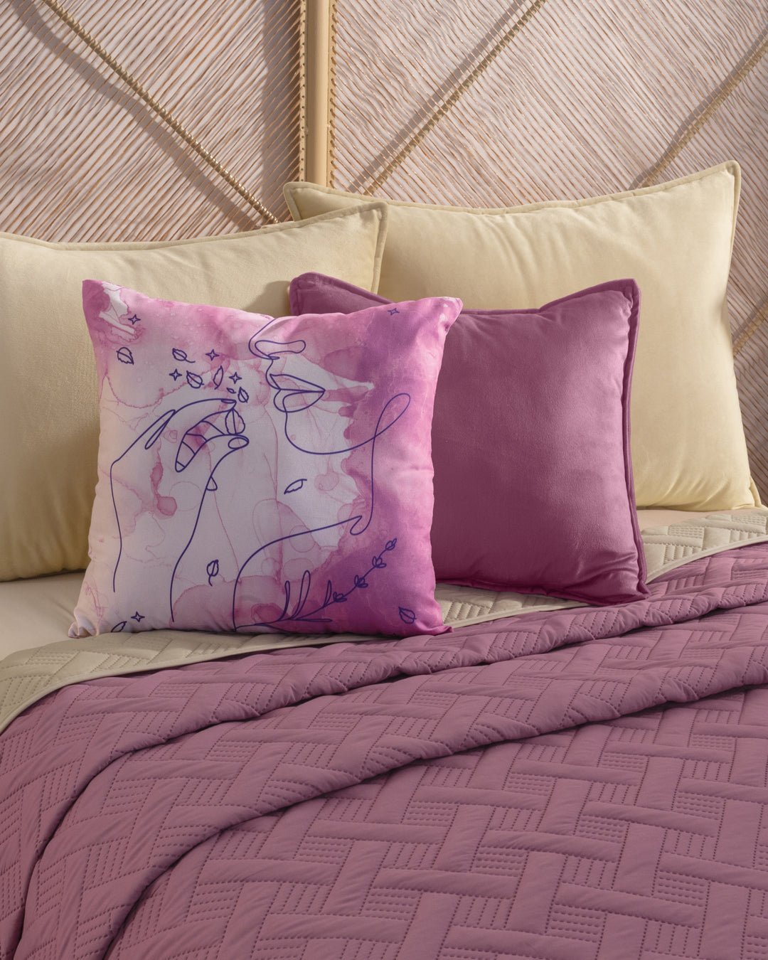 Two decorative throw pillows—one with pink abstract line art and one solid mauve—rest on a beige bed dressed in the Juego de Edredón Ligero Espíritu quilt by Intima Hogar, with beige pillows in the background.