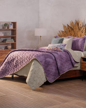 A neatly made bed features the Edredón Ligero Provenza by Intima Hogar in beige and purple, complemented by decorative pillows and a leaf-shaped wooden headboard. A nightstand and bookshelf complete this cozy, modern Provenza-inspired room.