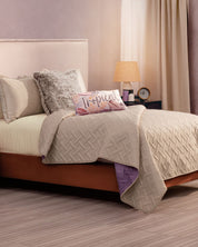 A cozy bed with Intima Hogar's Edredón Ligero Provenza in beige diseño reversible, a fluffy gray pillow, and a Tropical accent pillow. The bright room has light walls, curtains, and a bedside table with lamp, clock, and small decor.