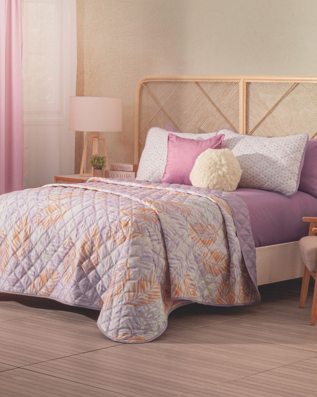 A cozy bedroom featuring the Intima Hogar Juego de Edredón Ligero Lima, lavender and white decorative shams, a fluffy round cushion, wooden headboard, bedside lamp, and soft pink curtains by the window.