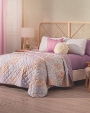 A cozy bedroom featuring the Intima Hogar Juego de Edredón Ligero Lima, lavender and white decorative shams, a fluffy round cushion, wooden headboard, bedside lamp, and soft pink curtains by the window.