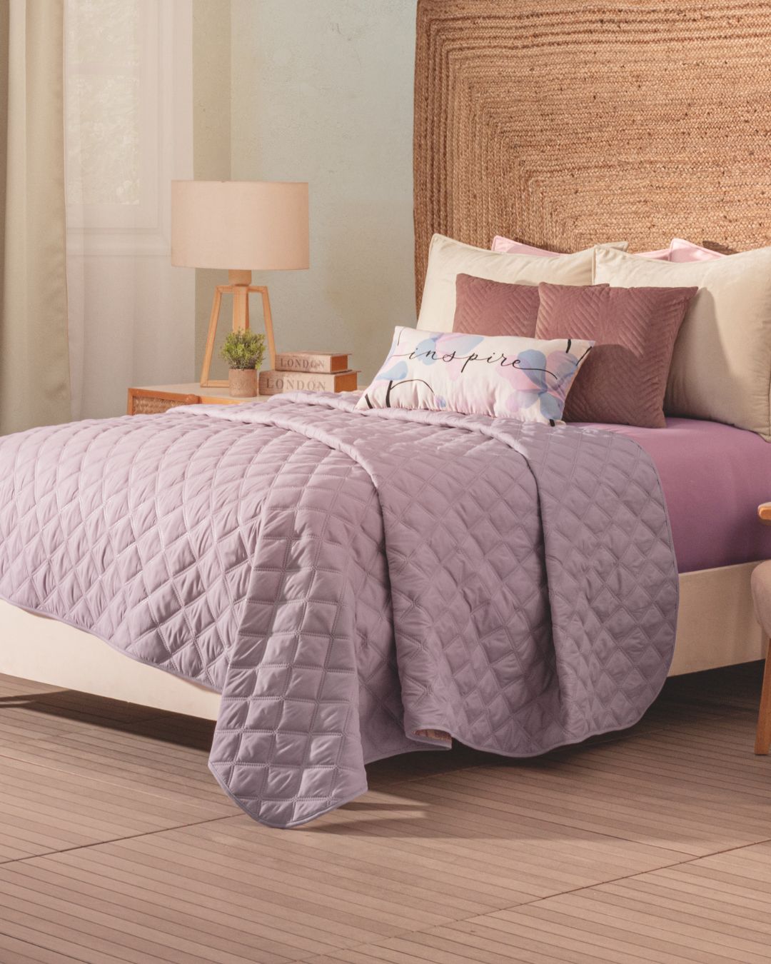 A neatly made bed features the Juego de Edredón Ligero Lima by Intima Hogar, beige and mauve decorative pillows, an inspire cushion, a woven headboard, and a nightstand with lamp and plant in a softly lit bedroom.