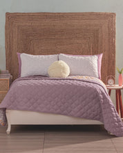 A neatly made bed with the Juego de Edredón Ligero Lima by Intima Hogar, two white patterned pillows, a round fluffy pillow, and a large woven headboard. Decorative covers and a small wooden nightstand with clock and plant complete the look.