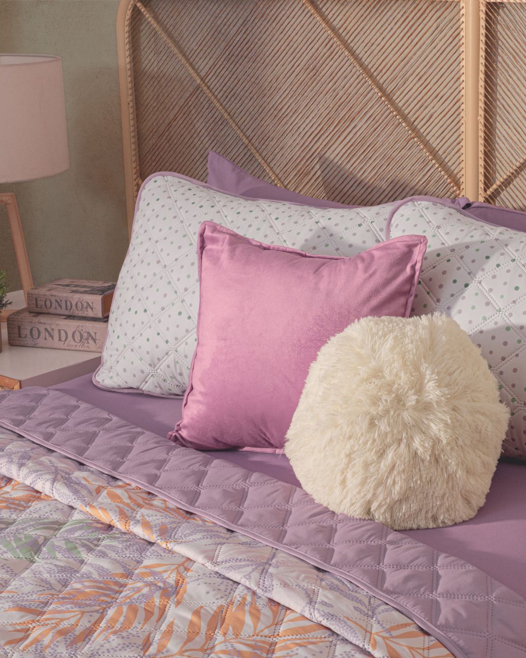 A bed is styled with the Juego de Edredón Ligero Lima by Intima Hogar, featuring lavender sheets, a pink square pillow, and a round fluffy white pillow. A woven headboard, books, and a lamp complete the cozy scene.