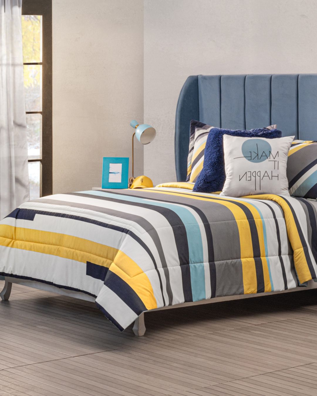 A neatly made bed features the Intima Hogar Juego de Edredón Luca in yellow, blue, gray, and white stripes, with decorative pillows including a "Make It Happen" case. A blue lamp and picture frame rest on the bedside table.