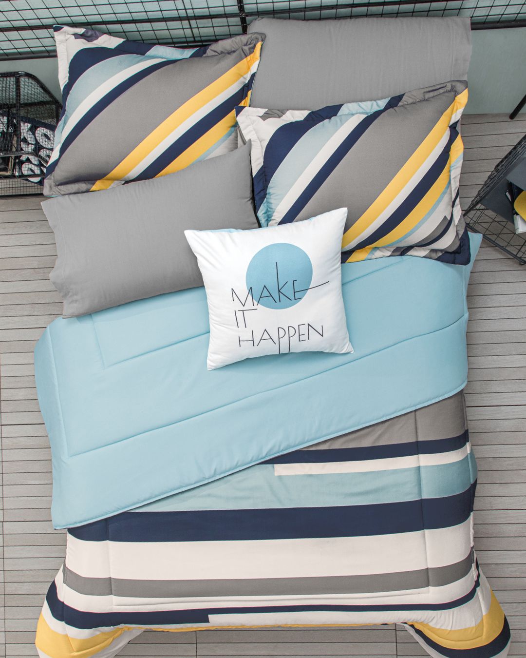 A neatly made bed features the Intima Hogar Juego de Edredón Luca with blue, yellow, gray, and white stripes, topped with a white decorative cushion reading MAKE IT HAPPEN in black. The bed is set on a light gray floor.