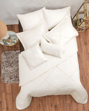 Overhead view of a neatly made bed with the Juego de Duvet Luxury Arena by Intima Hogar, featuring white bedding and pillows on a wooden floor beside a brown rug, a small glass table with lamp, and a gold shelf with decor.