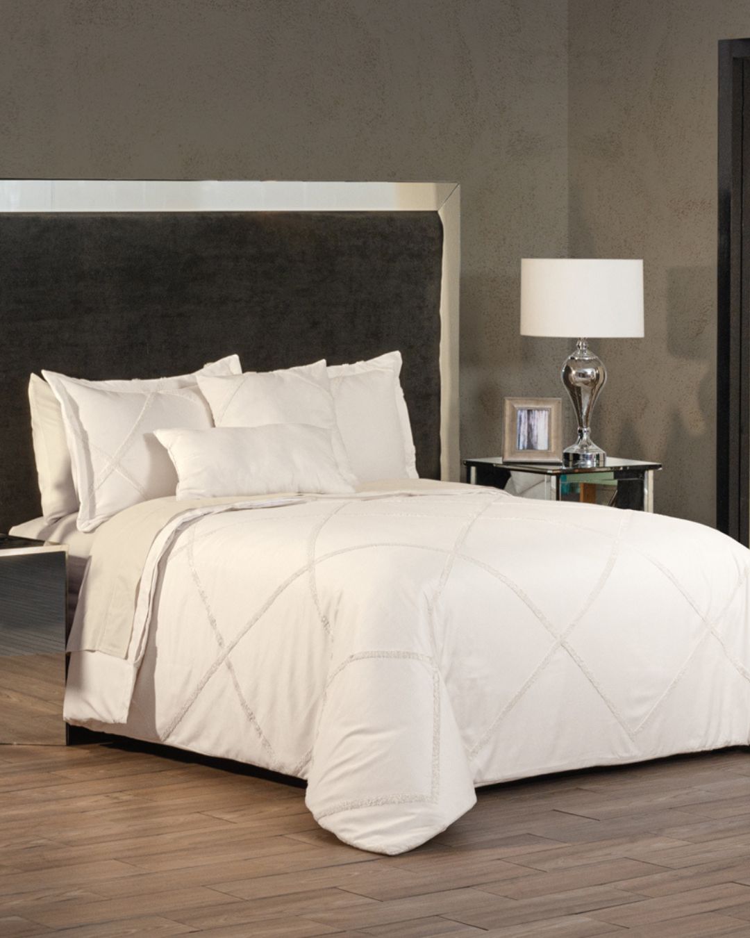 A neatly made bed features the Juego de Duvet Luxury Arena by Intima Hogar with several fundas decorativas against a dark upholstered headboard. A mirrored nightstand with a lamp and framed photo sits beside it, set against neutral-toned walls.