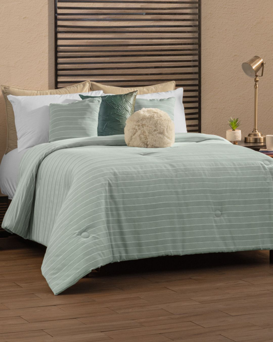 A neatly made bed with the Juego de Edredón Luxury Mantis by Intima Hogar, white pillows, and round green decorative cushions sits beside a small table with a lamp, plant, and books, set against a wooden headboard and beige wall.