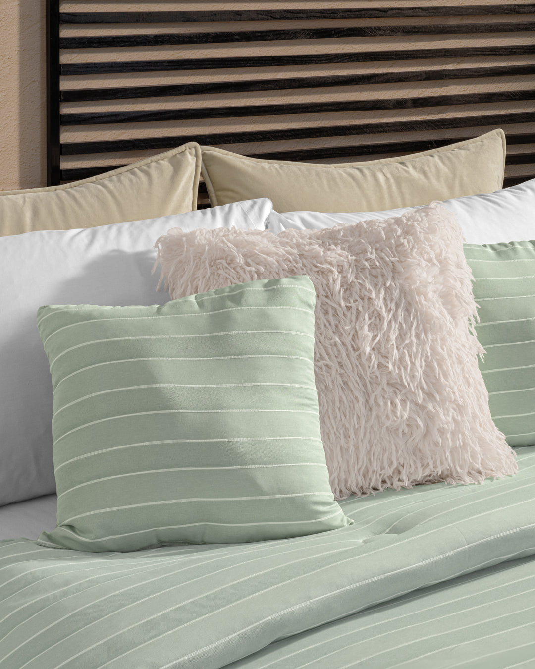 A neatly made bed features the Juego de Edredón Luxury Mantis by Intima Hogar, showcasing light green striped bedding and pillows, a white fluffy textured pillow, and beige pillows against a modern wooden slatted headboard.