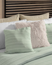 A neatly made bed features the Juego de Edredón Luxury Mantis by Intima Hogar, showcasing light green striped bedding and pillows, a white fluffy textured pillow, and beige pillows against a modern wooden slatted headboard.