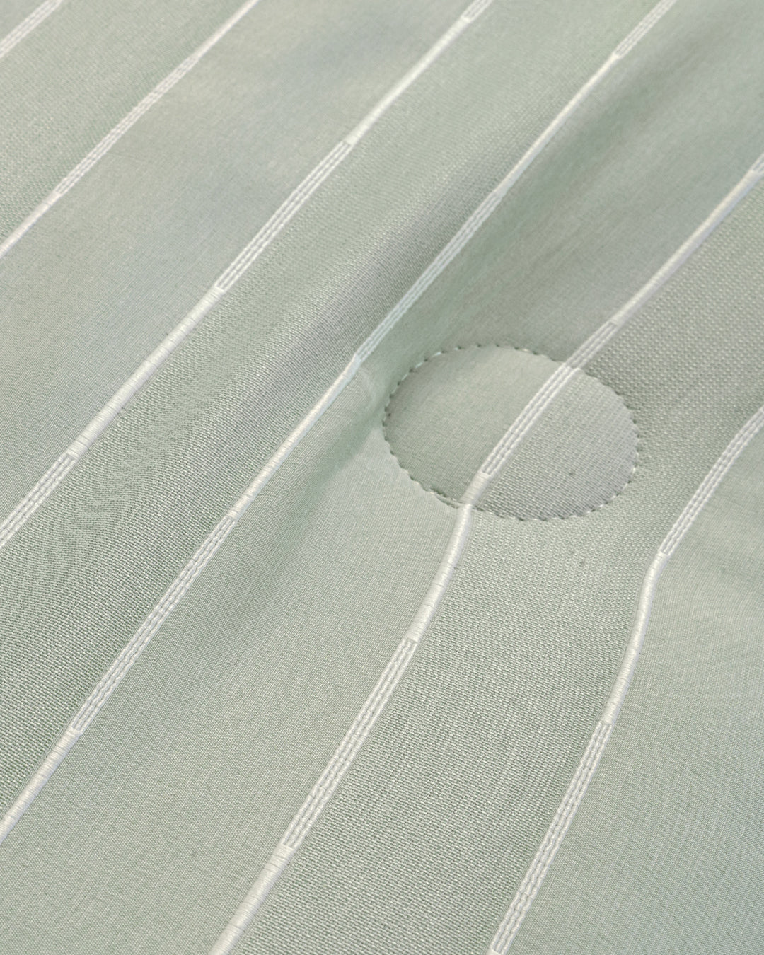 A close-up of the Juego de Edredón Luxury Mantis by Intima Hogar shows light green fabric with thin white vertical stripes and a stitched circular patch at the center right.