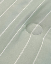 A close-up of the Juego de Edredón Luxury Mantis by Intima Hogar shows light green fabric with thin white vertical stripes and a stitched circular patch at the center right.