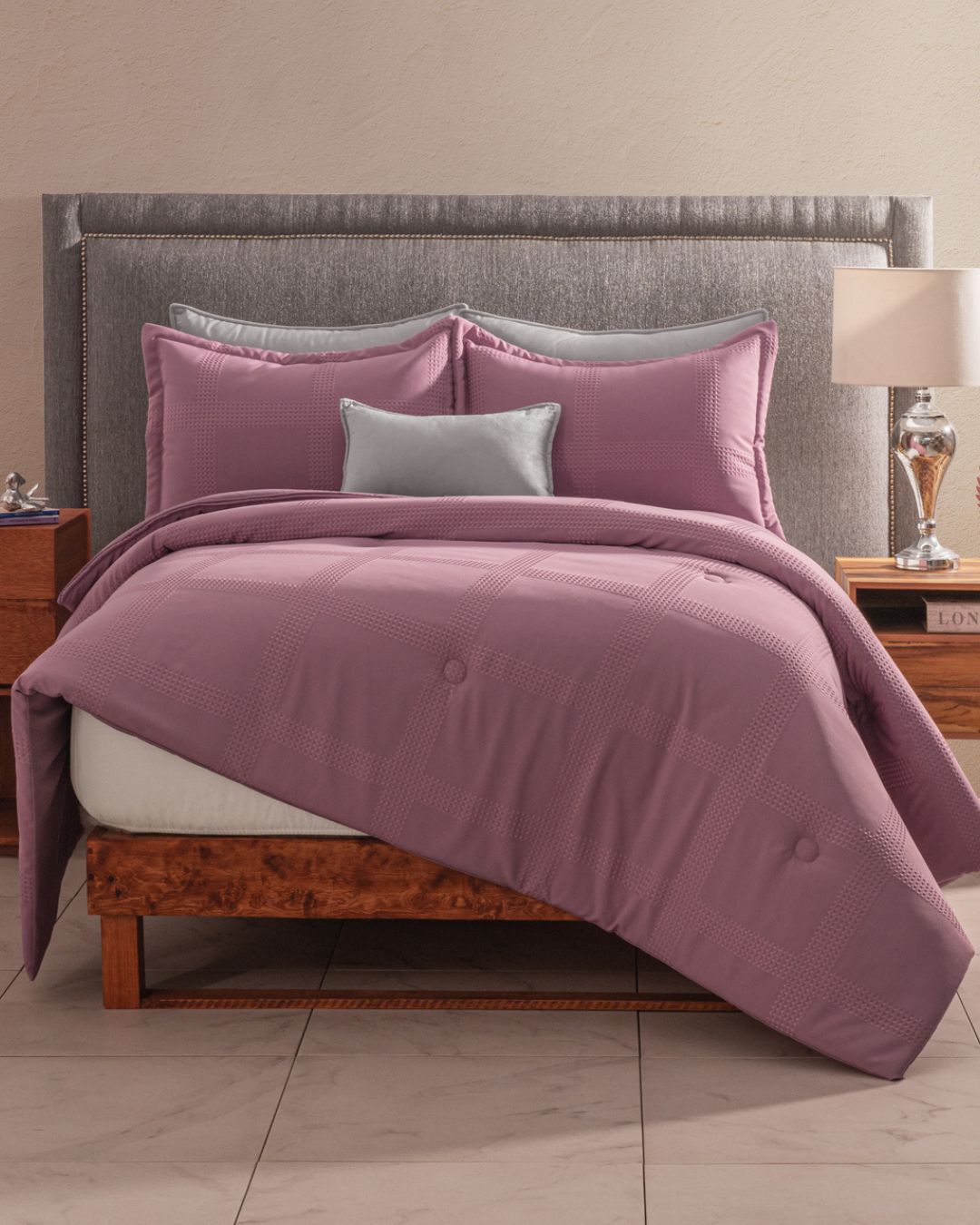 A neatly made bed features the Juego de Edredón Luxury Pétalo by Intima Hogar in mauve with matching pillows and a silver accent pillow. A light gray upholstered headboard and fundas decorativas complement the wooden nightstands with lamps.