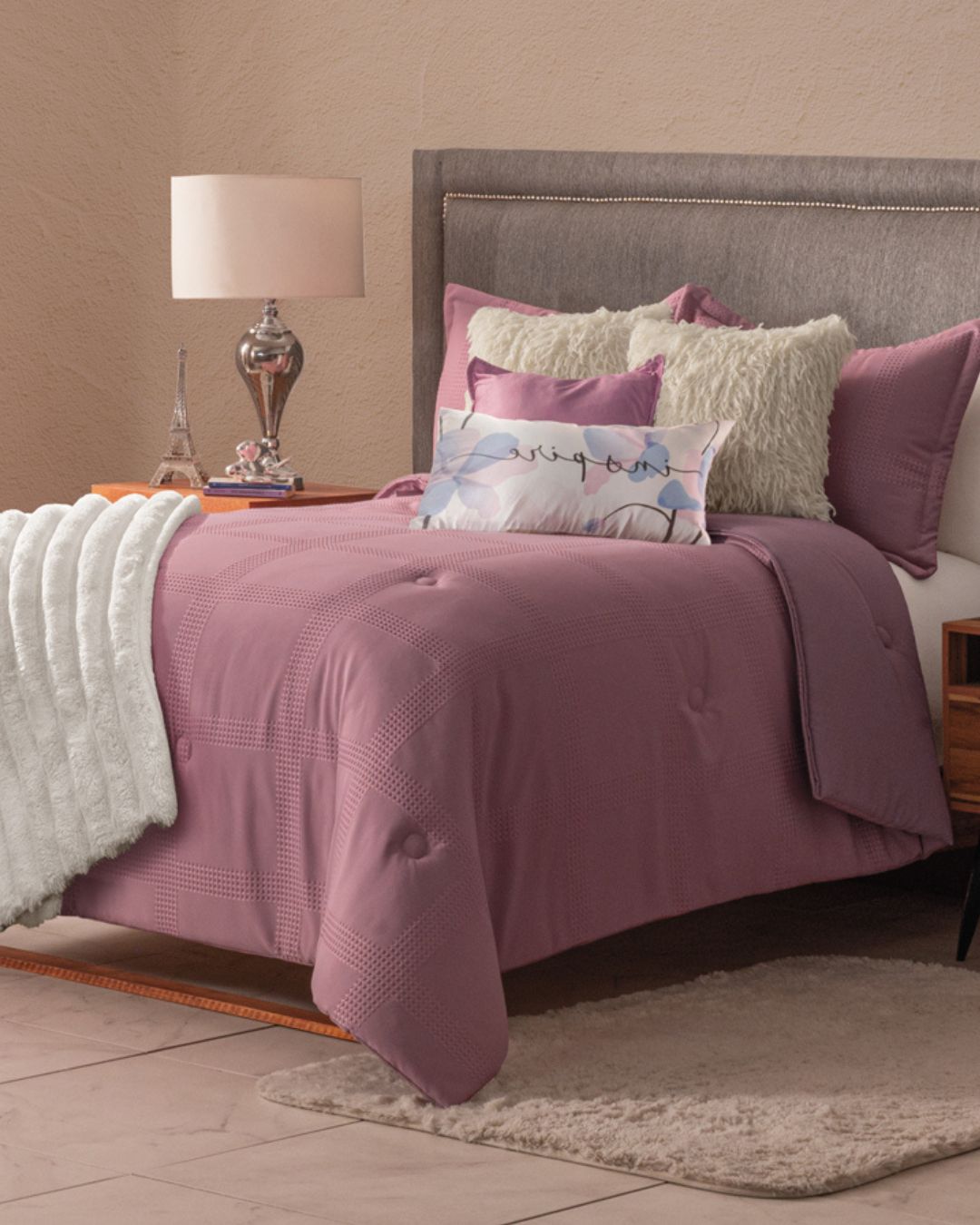 A neatly made bed features the Juego de Edredón Luxury Pétalo in mauve by Intima Hogar with fluffy white throws and decorative shams. A lamp and Eiffel Tower figurine sit on the bedside table, while a gray headboard completes the cozy decor.