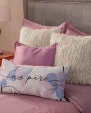 A bed is styled with the Juego de Edredón Luxury Pétalo by Intima Hogar, mauve bedding, white and mauve pillows, and a decorative pastel floral pillow reading "inspire". A lamp sits on the wooden nightstand beside the bed.