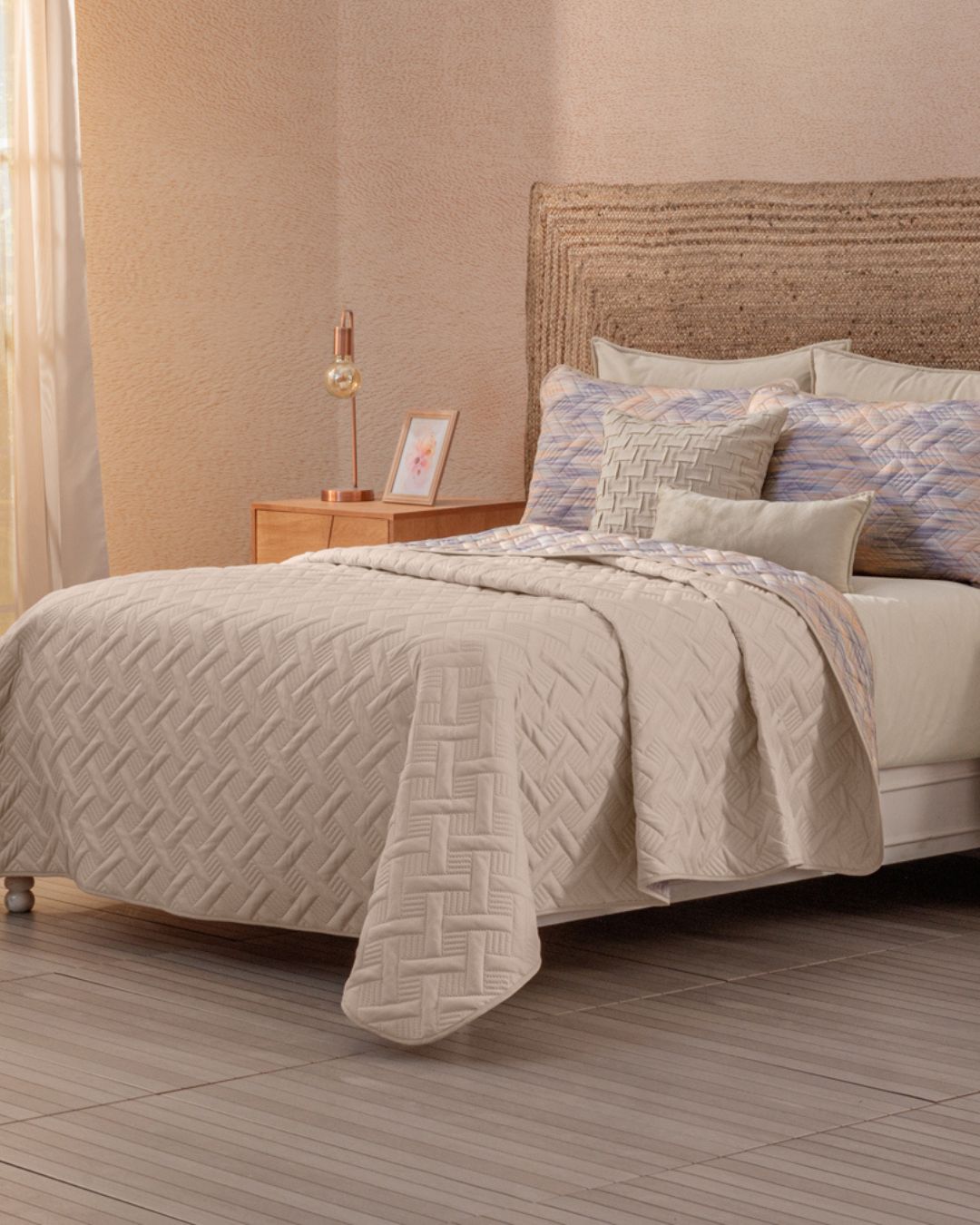 A neatly made bed features the Juego de Edredón Ligero Melodía by Intima Hogar, decorative pillows, a woven headboard, and a wooden nightstand with a lamp, picture frame, and book. Soft light enters through sheer curtains at the window.