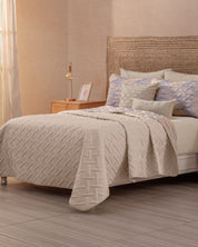 A neatly made bed features the Juego de Edredón Ligero Melodía by Intima Hogar, decorative pillows, a woven headboard, and a wooden nightstand with a lamp, picture frame, and book. Soft light enters through sheer curtains at the window.