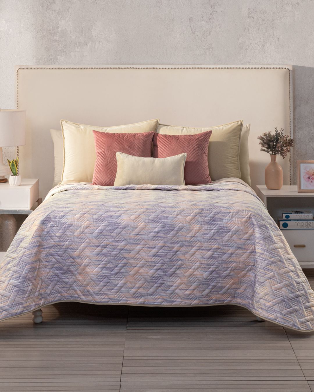 A neatly made bed features the Juego de Edredón Ligero Melodía by Intima Hogar, with its reversible design, cream and pink decorative covers, a cream headboard, and nightstands on each side topped with lamps and décor accents.