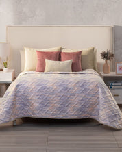 A neatly made bed features the Juego de Edredón Ligero Melodía by Intima Hogar, with its reversible design, cream and pink decorative covers, a cream headboard, and nightstands on each side topped with lamps and décor accents.