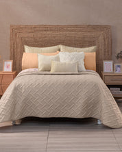 A neatly made bed features the Juego de Edredón Ligero Melodía by Intima Hogar in beige, with decorative shams, set against a woven headboard. Wooden nightstands with framed photos and decor rest on a light tile floor.