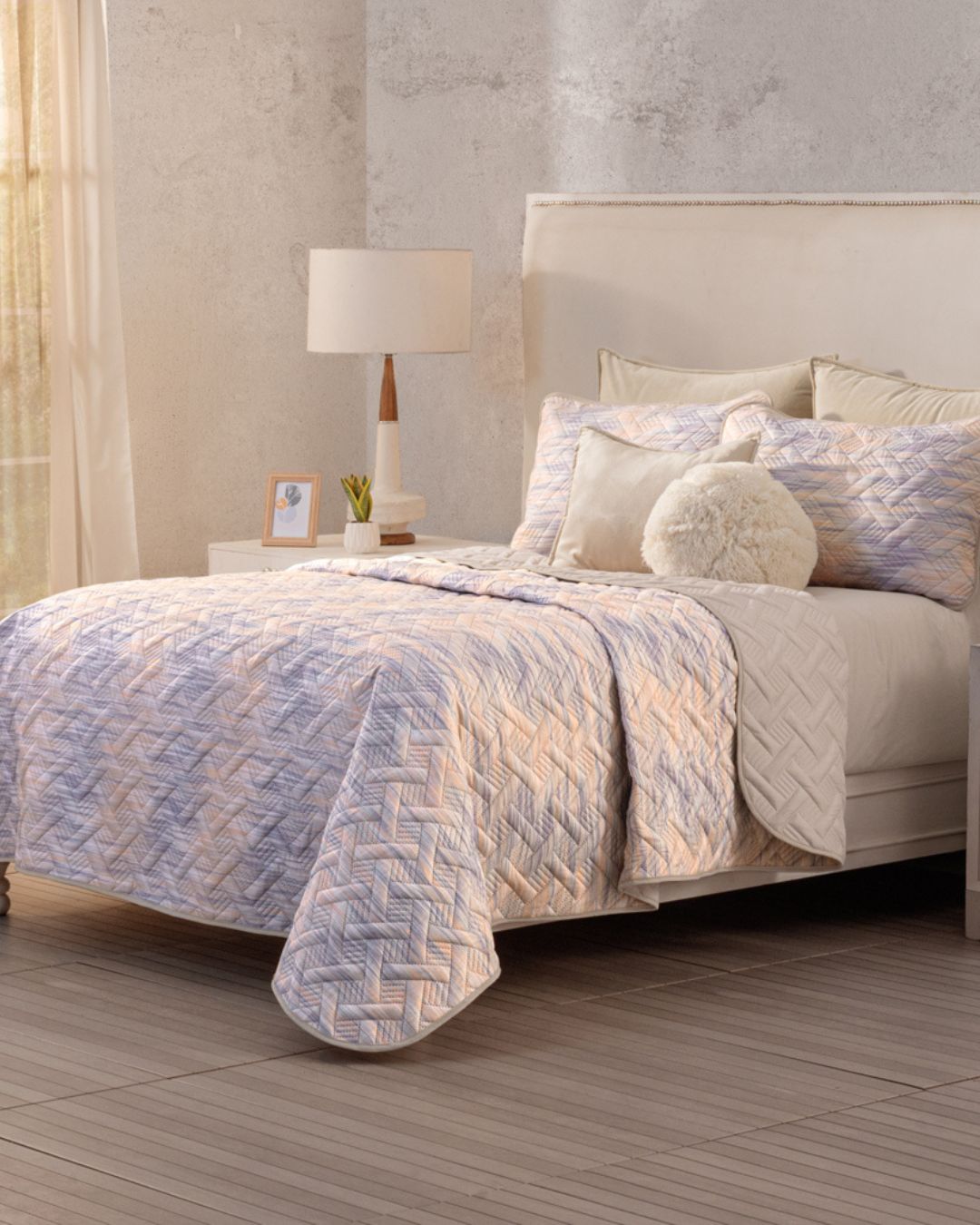 A neatly made bed features the Juego de Edredón Ligero Melodía by Intima Hogar, with matching pillows and fundas decorativas. A round fluffy cushion adds charm, while a bedside table, lamp, framed photo, and curtain let in natural light.
