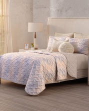 A neatly made bed features the Juego de Edredón Ligero Melodía by Intima Hogar, with matching pillows and fundas decorativas. A round fluffy cushion adds charm, while a bedside table, lamp, framed photo, and curtain let in natural light.