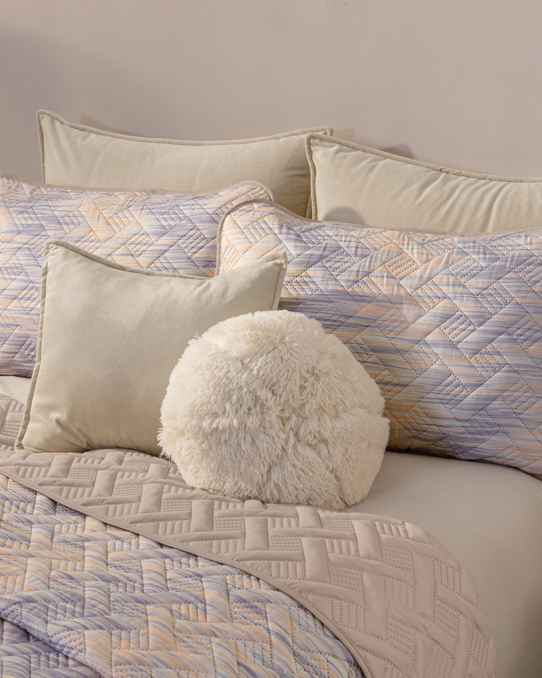 A neatly made bed features the Juego de Edredón Ligero Melodía by Intima Hogar, with quilted pastel bedding, geometric-patterned beige and lavender pillows, and a soft round white cushion in front.