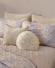 A neatly made bed features the Juego de Edredón Ligero Melodía by Intima Hogar, with quilted pastel bedding, geometric-patterned beige and lavender pillows, and a soft round white cushion in front.