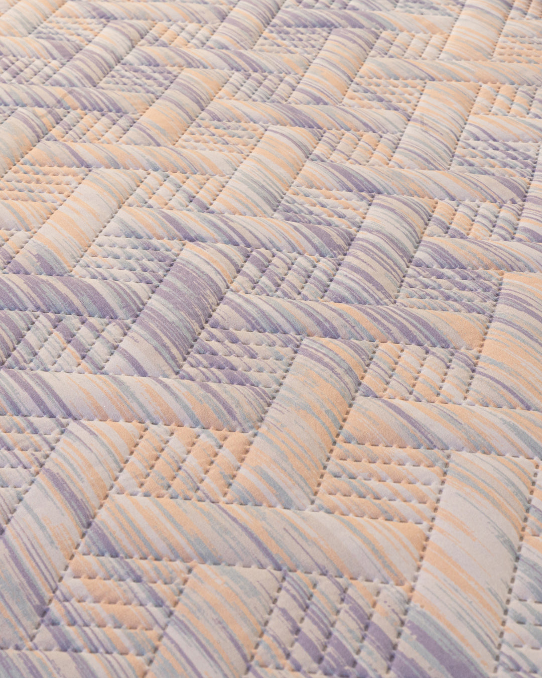 A close-up of Intima Hogar’s Juego de Edredón Ligero Melodía shows a woven, basket-like design in pastel lavender, peach, and cream tones with textured stitching and diagonal striped fabrics.