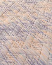 A close-up of Intima Hogar’s Juego de Edredón Ligero Melodía shows a woven, basket-like design in pastel lavender, peach, and cream tones with textured stitching and diagonal striped fabrics.