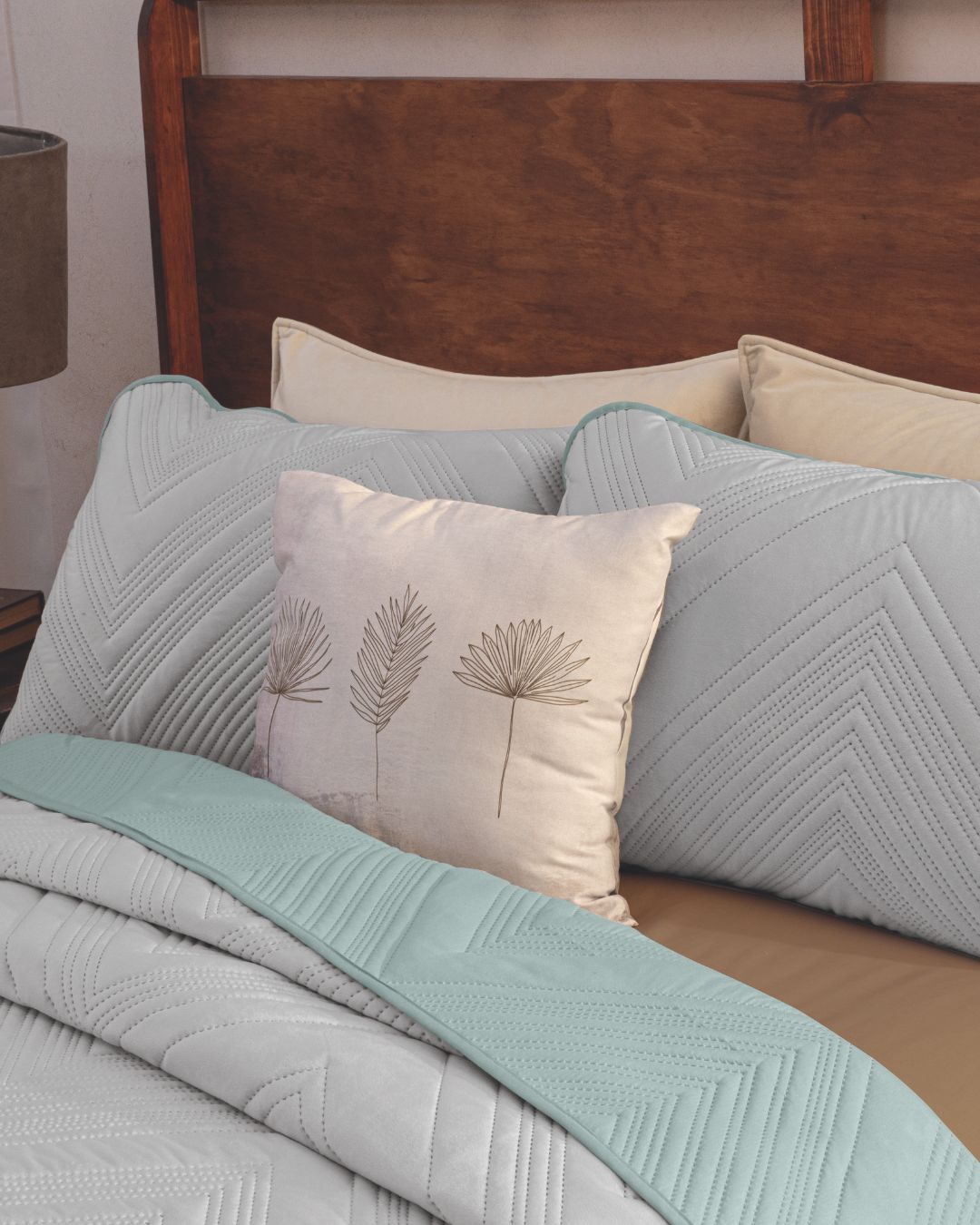 A bed styled with the Juego de Edredón Velvet Silencio by Intima Hogar features light gray quilted bedding, a soft mint blue throw, and decorative pillows—including a beige one with line-drawn botanical leaves—against a wooden headboard.