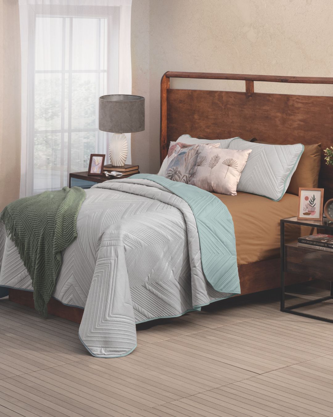 A neatly made bed features the Juego de Edredón Velvet Silencio by Intima Hogar in light blue and tan, paired with two leaf-pattern pillows and a green throw. A wooden headboard, nightstand, lamp, photo frame, and sheer curtains complete the scene.