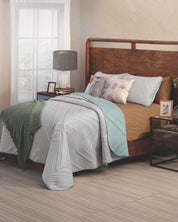 A neatly made bed features the Juego de Edredón Velvet Silencio by Intima Hogar in light blue and tan, paired with two leaf-pattern pillows and a green throw. A wooden headboard, nightstand, lamp, photo frame, and sheer curtains complete the scene.