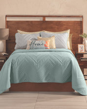A neatly made bed features the Intima Hogar Juego de Edredón Velvet Silencio, paired with cream and blue pillows and a decorative "Home" cushion. Two wooden nightstands with lamps and accents complete this restful setup.